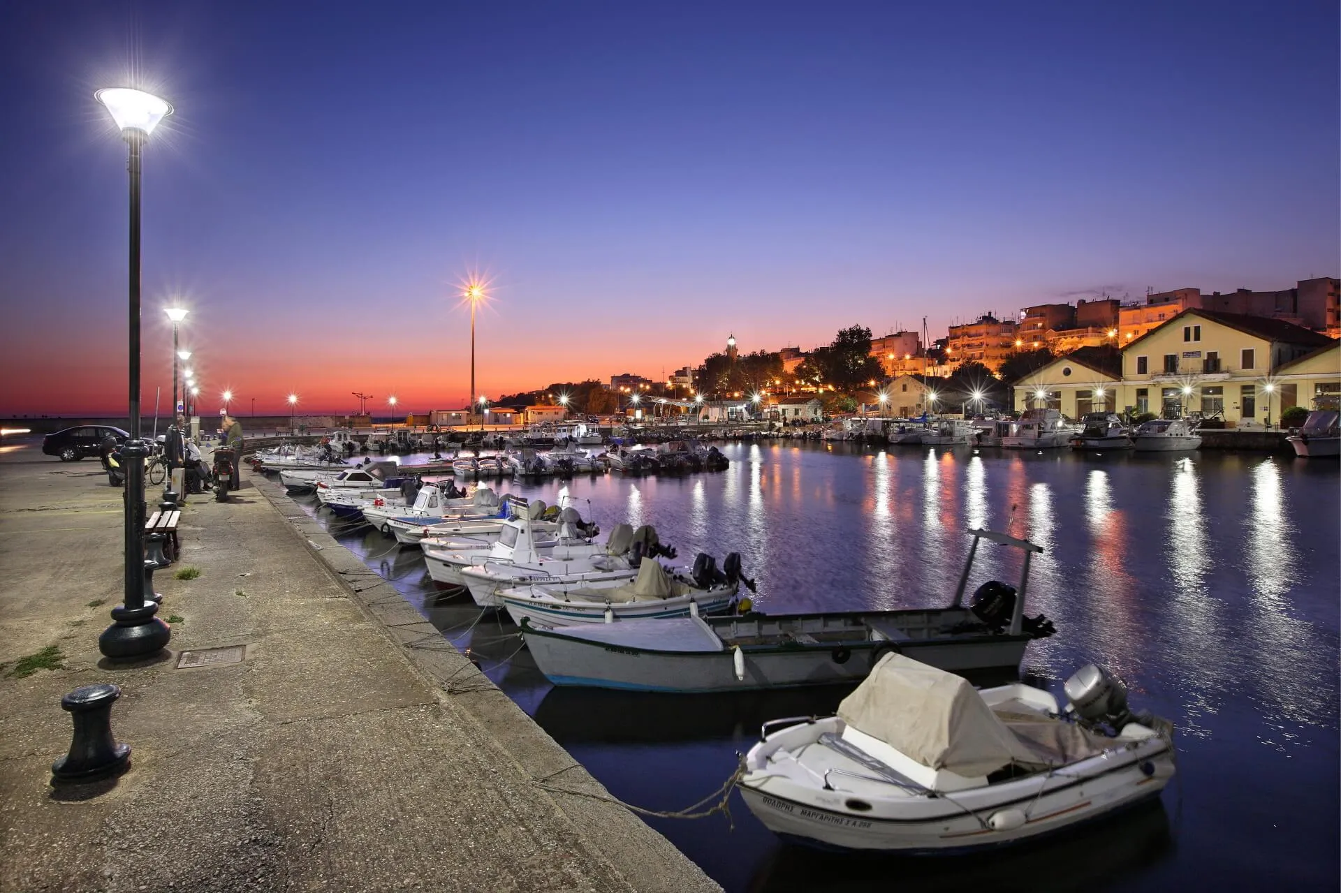 Fishing boats moored along the Alexandroupolis waterfront at dusk, with the lit promenade and old town behind